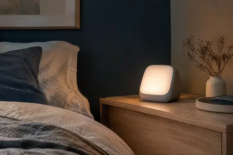 Réveil simulateur d’aube gris posé sur une table de chevet en bois dans une chambre tamisée.