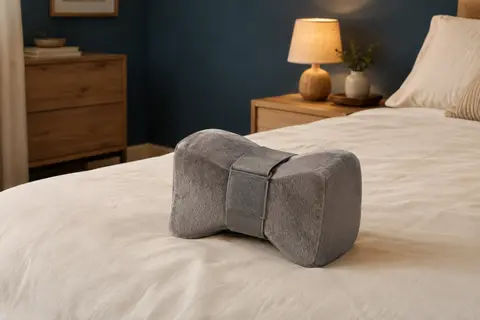 Coussin de genoux gris en mousse mémoire avec sangle réglable, posé sur un lit aux tons crème.