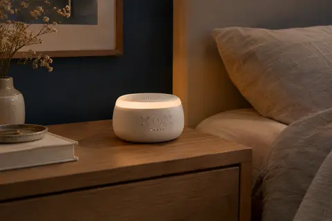 Machine à bruit blanc blanche avec veilleuse LED, posée sur une table de chevet en bois dans une chambre calme.
