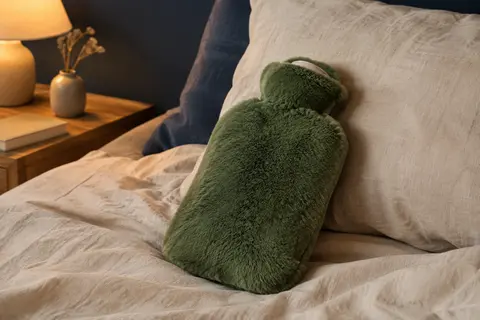 Bouillotte à eau avec housse en peluche verte posée sur un lit aux draps de lin crème