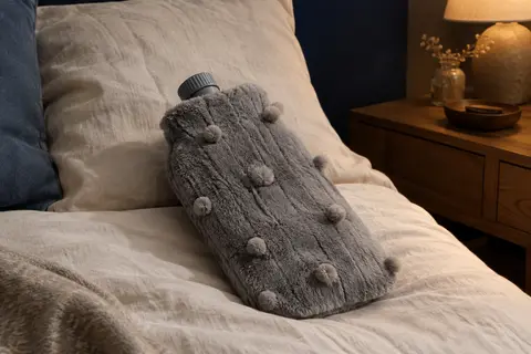 Bouillotte à eau nervurée avec housse grise douce en fausse fourrure posée sur un lit en lin.